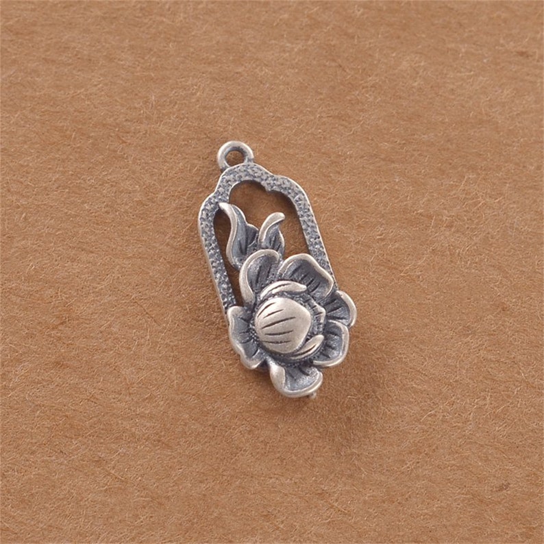 May include: A silver charm pendant featuring a detailed floral design. The flower is enclosed in a rectangular frame with an ornate border.