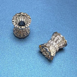 May include: Two silver-toned jewellery pieces with intricate designs. One piece is a small cup-shaped ring with a clear stone in the centre. The other piece is a bow-shaped ring with clear stones.