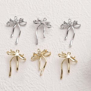 May include: Six bow-shaped charms in silver and gold tones. Each charm features a bow design with dangling ribbons and a small loop at the top for attachment. Some bows have a small, clear gem in the center.