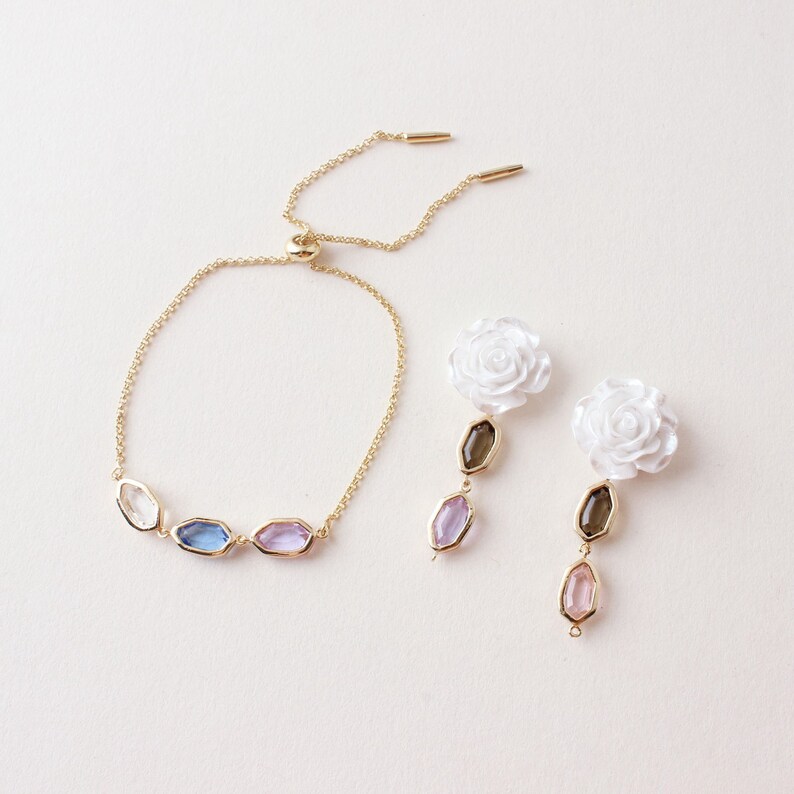 May include: A gold-toned bracelet with five colorful, faceted stones and a pair of earrings. The earrings feature white rose-shaped studs, a brown stone, and a pink stone. The jewelry is set against a white background.