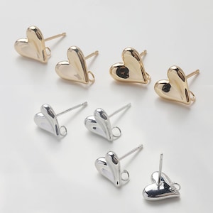 Pode incluir: Uma coleção de brincos de coração em ouro e prata. Os brincos têm um acabamento liso e polido e um pino com um pequeno laço na base do coração. Os brincos dourados têm uma aparência quente e brilhante.