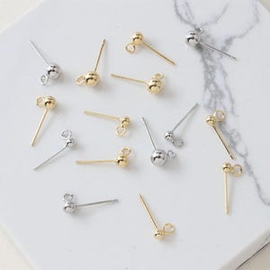 May include: A collection of gold-toned and silver-toned metal earring posts featuring a small spherical ball top and an attached open jump ring loop at the base for jewelry making, scattered across a white surface with grey marble veining.