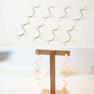 May include: A collection of eight silver and gold-toned metal spiral earring components, along with a pair of assembled gold-toned spiral earrings featuring small white beads and a geometric cube charm displayed on a wooden T-bar stand.