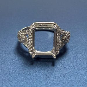 Puede incluir: Un engaste de anillo de plata con una piedra central de talla esmeralda cuadrada y diamantes pavé en la banda.
