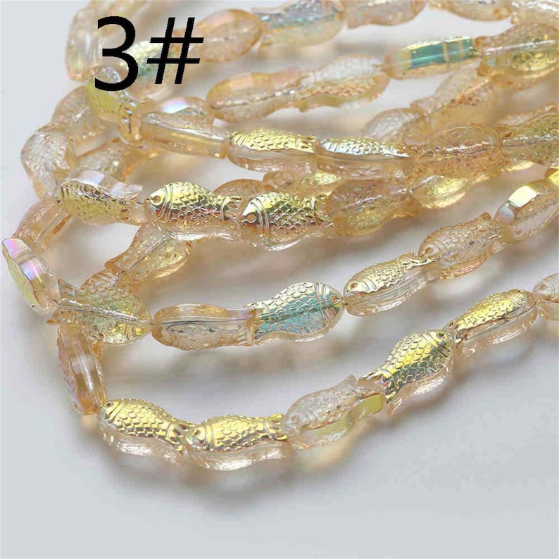 May include: A strand of iridescent glass beads shaped like fish. The beads are a mix of light gold and clear glass with a subtle shimmer. The beads are strung on a clear thread. The image includes the text "3#" at the top.