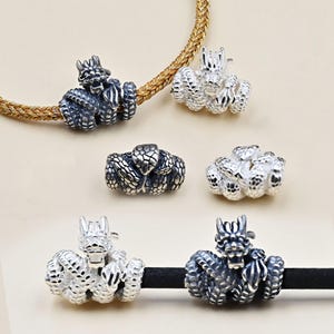 May include: Silver dragon charm beads in various styles. The charms are detailed with scales and horns, and are shown on a gold chain and a black cord. The beads are in different shades of silver, with some appearing darker than others.