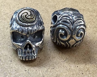 Puede incluir: Dos cuentas de plata en forma de calavera, una con un diseño de espiral dorado en la parte superior y la otra con un patrón de espiral.