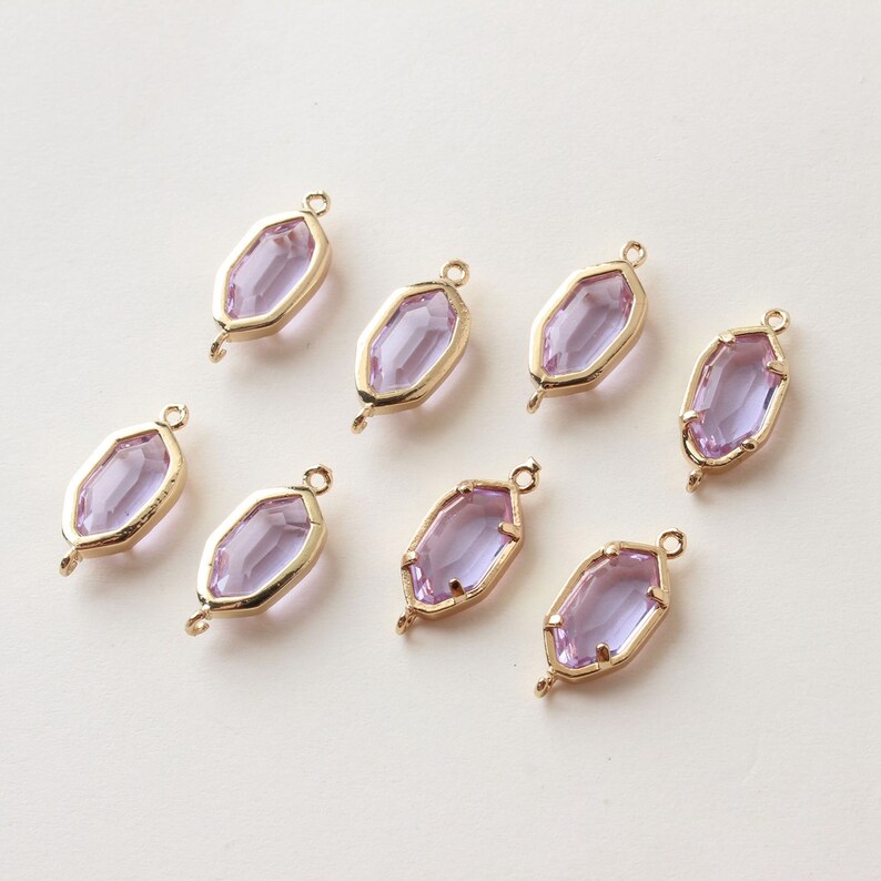 May include: Eight lavender-colored, faceted glass charms with gold-colored metal frames. Each charm has a hexagonal shape with a loop at the top and bottom for attaching to jewelry. The charms are arranged on a light-colored surface.