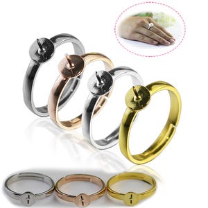 May include: A collection of adjustable ring blanks in silver, rose gold, silver, and gold. Each ring features a flat, round setting with a central post for attaching beads or other embellishments. The rings are displayed on a white surface.