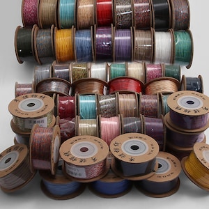 Puede incluir: Una colección de numerosas bobinas de cordón de 1,0 mm en una gran variedad de colores sólidos y patrones multicolores. Cada bobina presenta una tapa circular de cartón con caracteres chinos impresos y el texto 1,0 mm, dispuestas en pila.