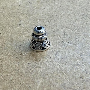 Puede incluir: Una pequeña cuenta de color plateado con un diseño escalonado. La parte superior presenta un agujero, seguido de una sección acanalada y una base con símbolos grabados. La cuenta probablemente se utiliza para joyería o artesanía.