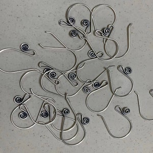 Puede incluir: Una colección de ganchos para pendientes de color plateado con un diseño de espiral decorativo. Los ganchos están hechos de alambre fino y tienen una apariencia simple y elegante. El detalle de la espiral es de un color más oscuro, agregando contraste.