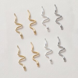 Może przedstawiać: Sześć złotych i srebrnych zawieszek w kształcie węża z pętelką na końcu. Zawieszki są ułożone w dwóch rzędach po trzy.