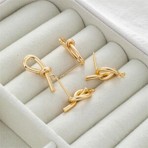 May include: Four gold-colored earrings with a knot design are displayed in a white jewelry box. Two are stud earrings, and two are dangle earrings. The earrings are made of a shiny, metallic material.