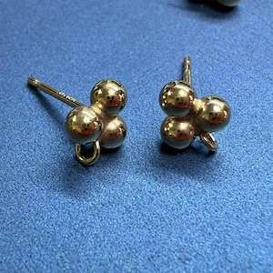 May include: A pair of gold-colored stud earrings. Each earring features a cluster of three small, round spheres. The earrings have a post and a curved backing for secure wear. The background is a solid blue.