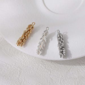 Puede incluir: Tres colgantes metálicos en forma de espiga de trigo, en oro, plata y un tono plateado más oscuro. Cada colgante tiene un pequeño lazo en la parte superior para sujetarlo a la joyería. Los colgantes están dispuestos sobre una superficie blanca.