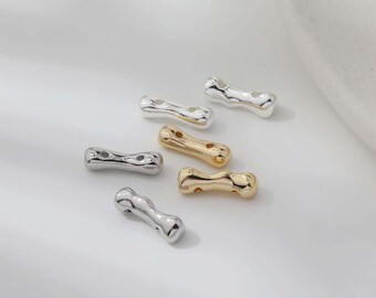 May include: An assortment of metallic beads in silver and gold tones. The beads have a unique, elongated shape with two holes for stringing. The shiny beads are scattered on a white surface, creating a visually appealing arrangement. Ideal for jewelry making.