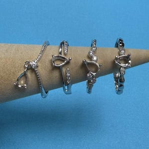 May include: Four silver rings with clear stones. The rings are on a wooden display stand. The rings are all different styles, but they all have a similar design.