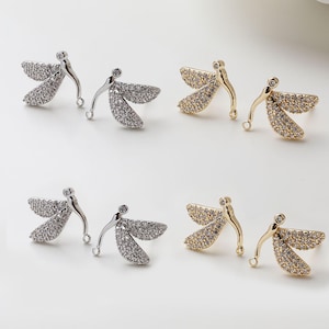 Op de afbeelding: Close-up van twee paar libellen-oorstekers. Eén paar is zilverkleurig met heldere stenen, de andere is goudkleurig met heldere stenen. De oorbellen hebben een gebogen lichaam en gedetailleerde vleugels.