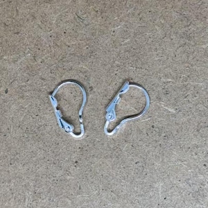Könnte beinhalten: Zwei silberne Ohrstecker, auch Ohrbügel genannt, sind auf einer braunen Oberfläche zu sehen. Die Ohrstecker sind aus Metall und haben einen federbelasteten Verschluss.