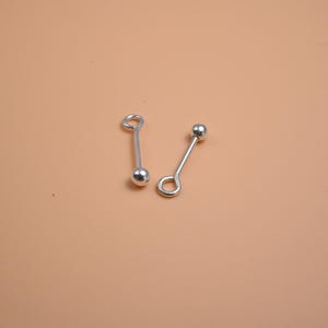 May include: Two silver-colored tie tacks with a ball at one end and a loop at the other. The tie tacks are displayed on a light brown surface. The tie tacks are small and delicate.
