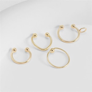 May include: A collection of gold-colored, horseshoe-shaped nose rings and a circular ring. Each ring features a small ball at the ends. One ring has a small loop attached. The jewelry is displayed on a white surface.