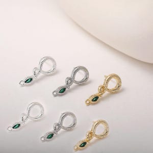 Könnte beinhalten: Nahaufnahme mehrerer schlangenförmiger Schmuckanhänger. Die Anhänger sind silber- und goldfarben und haben eine strukturierte Oberfläche. Jeder Anhänger hat einen grünen, oval geformten Edelstein. Die Anhänger sind für die Schmuckherstellung bestimmt.