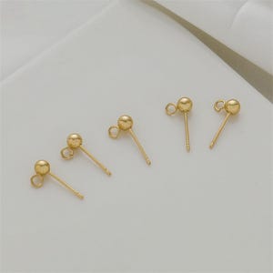 May include: Five gold-colored earring posts with a small ball at the top and a loop for attaching charms. The posts are arranged diagonally on a white surface. The earrings are simple and elegant.