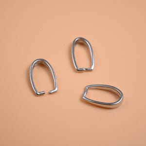 Puede incluir: Tres fornituras de joyería plateadas, cada una con una forma ovalada abierta única. Las fornituras se muestran sobre un fondo de color melocotón sólido. Estas fornituras probablemente se utilizan en la fabricación de joyas.