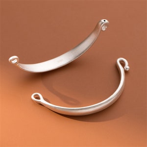 Könnte beinhalten: Zwei silberfarbene Metall-Armbandverschlüsse mit einer gebogenen Form und einer kleinen Schlaufe an jedem Ende.