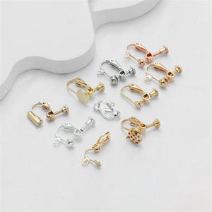 May include: A collection of clip-on earring findings in various metallic finishes, including gold, silver, and rose gold. The findings feature different designs, including screw-back and lever-back styles, and are displayed on a white surface.