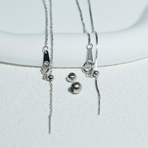 May include: Silver chain earrings featuring small, dangling silver beads and a bar at the end. Two additional silver beads are also present. The earrings are displayed on a white surface.
