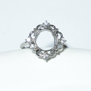 May include: A silver ring with an ornate, openwork design. The ring features a central oval setting surrounded by decorative flourishes and small, clear gemstones. The ring is set against a white background.