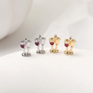 May include: Miniature champagne glass charms in silver and gold tones, each with a heart-shaped red gem. The charms are arranged in pairs, suggesting a theme of celebration or romance. The design is delicate and detailed.