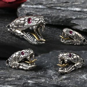 May include: Four silver-toned metal snake head beads with textured scales, open mouths revealing gold-toned tongues, and small red circular stones set as eyes. Each bead features a detailed reptilian design with sharp fangs and a metallic finish.
