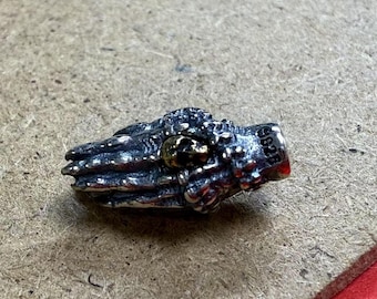 Puede incluir: Un amuleto de plata y oro en forma de manos en oración con un detalle de calavera. El amuleto está marcado con el número "925".