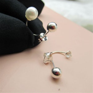 May include: A pair of silver earrings with a unique design. One earring features a large, lustrous white pearl, while the other has a small, shiny silver ball. The earrings are displayed against a black fabric and a light pink background.