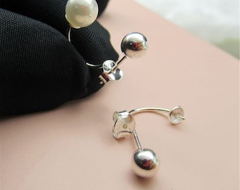 May include: A pair of silver earrings with a unique design. One earring features a large, lustrous white pearl, while the other has a small, shiny silver ball. The earrings are displayed against a black fabric and a light pink background.