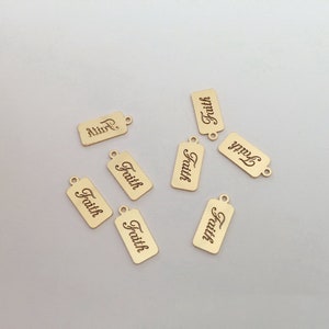 Pode incluir: Pingentes retangulares dourados com a palavra "Faith" gravada em fonte cursiva. Cada pingente tem um pequeno orifício na parte superior para prender a joias. Os pingentes estão espalhados em uma superfície de cor clara.