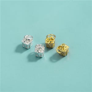Puede incluir: Cuatro pequeños dijes de metal con forma de cajas de regalo, cada uno rematado con una argolla circular. Dos de los dijes tienen un acabado plateado brillante y dos tienen un acabado dorado intenso, dispuestos en un grupo suelto.