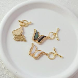 May include: Three gold-toned toggle clasps featuring butterfly-shaped charms. One charm has a textured gold finish, one has dark iridescent wings, and one has white iridescent wings. Each butterfly is accented with small clear stones along the edges.
