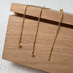 Könnte beinhalten: Drei goldfarbene Ohrringe mit filigranen Ketten und kleinen, runden Stiften. Ein Ohrring hat ein gerades Stangendesign. Die Ohrringe werden auf einer Holzoberfläche präsentiert, was ihre elegante Schlichtheit hervorhebt.