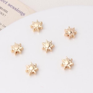 May include: Six small, gold-colored sun-shaped charms. Each charm has a circular center with rays extending outwards. The charms are scattered on a white surface, suggesting they are jewelry components or decorative elements.