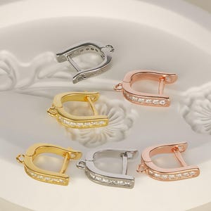 May include: An assortment of hoop earrings in silver, gold, and rose gold tones. Each earring features a row of small, clear gemstones along the curved edge. The earrings are displayed on a white surface.