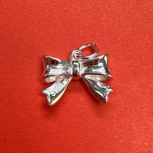 May include: A silver charm in the shape of a bow. The charm has a loop at the top for attaching to a bracelet or necklace. The charm is marked with the number "925" on the back, indicating that it is made of sterling silver.