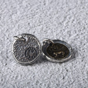 May include: Two silver-toned pendants with a textured surface. One pendant features a stylised design of a vase with a crown on top. The other pendant features a stylised design of a lion with wings.