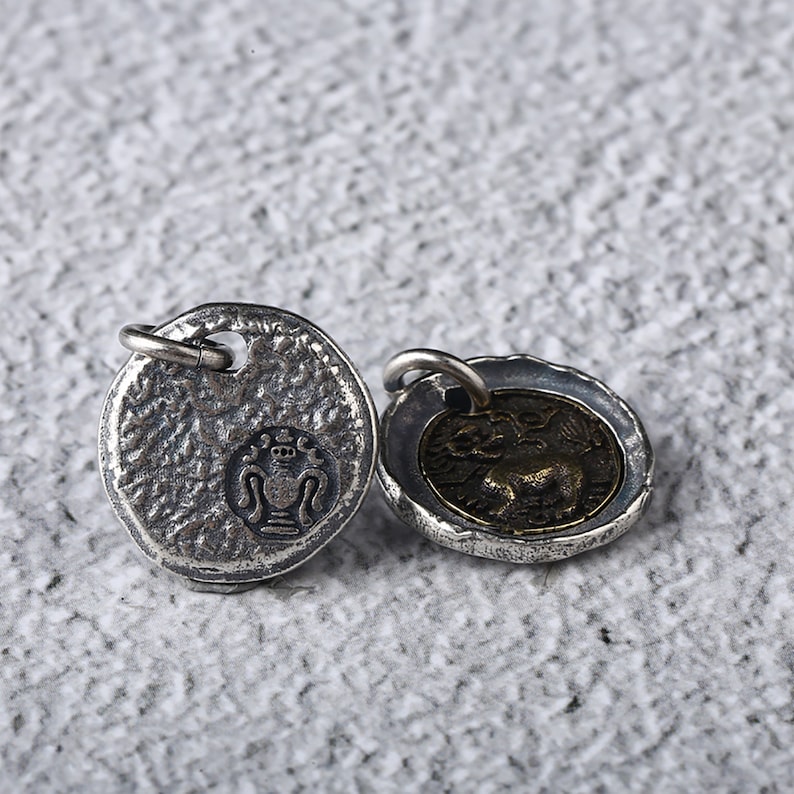 May include: Two silver-toned pendants with a textured surface. One pendant features a stylised design of a vase with a crown on top. The other pendant features a stylised design of a lion with wings.