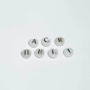 Può includere: Sette perline bianche e rotonde con lettere nere che compongono "HEAL CR I". Le perline sono disposte a semicerchio su sfondo bianco. Le lettere sono in un semplice font sans-serif.
