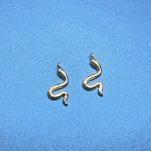 May include: Two gold-coloured snake-shaped pendants against a blue background. The pendants have a curved design, and each has a small loop at the top for hanging. The jewellery is likely for necklaces or earrings.