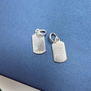 Puede incluir: Dos dijes de plata en forma de placa de identificación de perro con una superficie lisa ligeramente texturizada. Los dijes tienen un anillo en la parte superior para sujetarlos a un collar o pulsera. Uno de los dijes tiene la inscripción "S925" en la parte delantera.